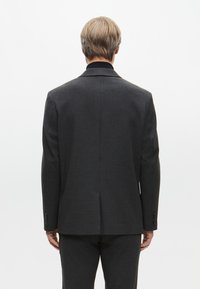 Giacca da abito grigia su misura con rever a tromba, maniche lunghe e uno spacco singolo sul retro. Il tessuto appare strutturato con un sottile motivo.