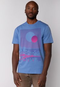 Blauw T-shirt met korte mouwen, voorzien van een abstract grafisch ontwerp met een grote blauwe cirkel en een roze cirkel, tegen een achtergrond van gestreepte patronen.