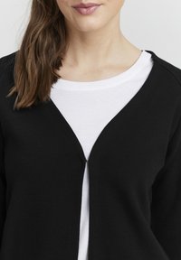 Černý žebrovaný cardigan s výstřihem do V, volným předním zapínáním a texturovaným povrchem, nošený přes bílou košili s kulatým výstřihem.