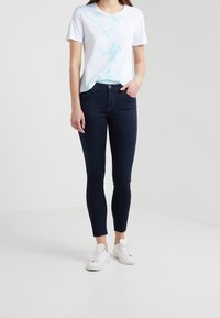 Mörkblå slim fit-jeans med hög midja och femfickorsdesign, kombinerade med en kortärmad vit t-shirt med ljusblå tie-dye-accenter.