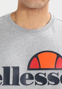 Szara bluza z okrągłym dekoltem, teksturowaną tkaniną oraz dużym, pomarańczowo-czarnym graficznym logo na przodzie.