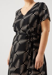 Zwarte jurk met een V-hals en korte mouwen, voorzien van een beige geometrisch bladdessin en een strikdetail in de taille, gemaakt van een luchtige stof.