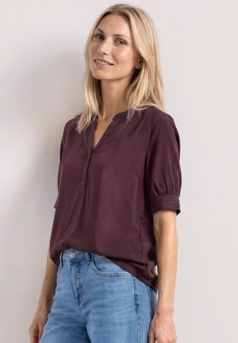 Femme blonde souriant légèrement, portant un chemisier à manches courtes violet foncé et un jean bleu clair, debout devant un fond clair uni.