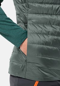 Gilet verde imbottito con sezioni quiltate orizzontali. Presenta una tasca laterale ed è indossato sopra una maglietta a manica lunga di colore teal.