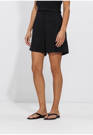 Vrouw die zwarte shorts met hoge taille en zwarte platte sandalen draagt, staand tegen een effen witte achtergrond.