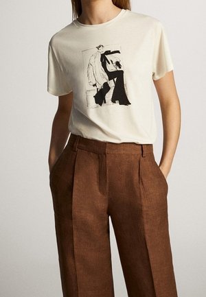 Woman wearing a cream t-shirt with a black and white graphic and high-waisted brown trousers with hands in pockets.