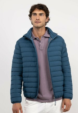 Hombre con chaqueta acolchada azul sobre una camisa polo morada y pantalones blancos, mirando ligeramente hacia la derecha sobre un fondo liso.