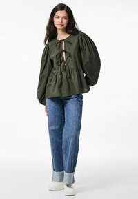 Blusa oliva con maniche a sbuffo, scollo profondo a V e dettagli con fiocco, abbinata a jeans di denim azzurro chiaro e sneakers bianche.