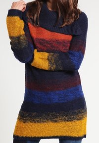 Pull en tricot avec col châle, présentant des rayures horizontales en bleu marine, rouge, orange, jaune et bleu, avec une texture douce.