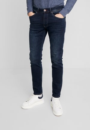 Pantalones vaqueros de mezclilla azul oscuro con un corte ajustado, que cuentan con bolsillos frontales y cierre de botón, que se combinan con zapatillas blancas.