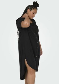 Robe noire à manches courtes, en tissu doux, avec une fente sur le côté et une coupe décontractée. Accentuée par des boutons dorés sur le devant.