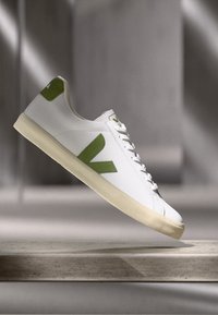 Hvit sneaker med grønne detaljer, med en teksturert gummisåle, snøring og en fremtredende logo på siden. Minimalistisk design.