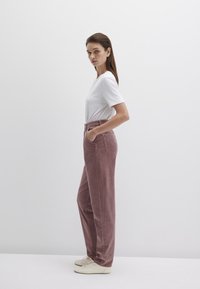 Hoch geschnittene Kordhosen in staubigem Rosa, mit seitlichen Taschen und einer lockeren Passform. Kombiniert mit einem einfachen weißen T-Shirt und hellen Sneakers.