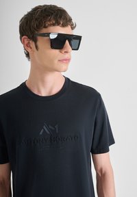 Camiseta negra con un destacado logotipo de la marca en relieve en el centro. El modelo lleva unas gafas de sol negras de gran tamaño con un diseño geométrico.