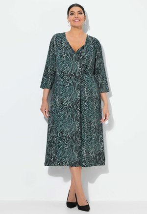 Langes Wickelkleid mit einem turquoise-schwarzen Schlangenprint-Muster, Dreiviertelärmeln und einem V-Ausschnitt. Schwarze High Heels ergänzen das Outfit.