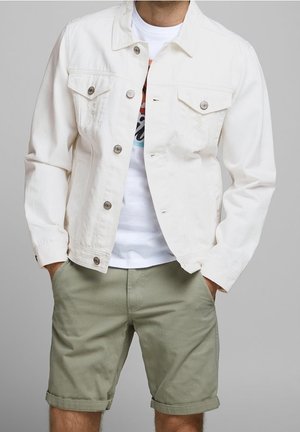 Homme portant une veste blanche boutonnée sur un t-shirt blanc graphique et un short olive clair retroussé avec les mains dans les poches.