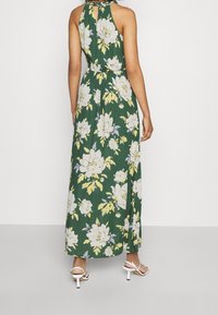 Kvinna i grön halterneck maxi-klänning med stort blommönster i vitt och gult samt vita klacksandaler, stående mot en enfärgad bakgrund.