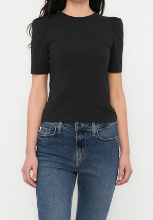 Vrouw draagt een zwart topje met korte mouwen en geplooide schouders, gecombineerd met een high-waisted blauwe spijkerbroek, tegen een effen witte achtergrond.