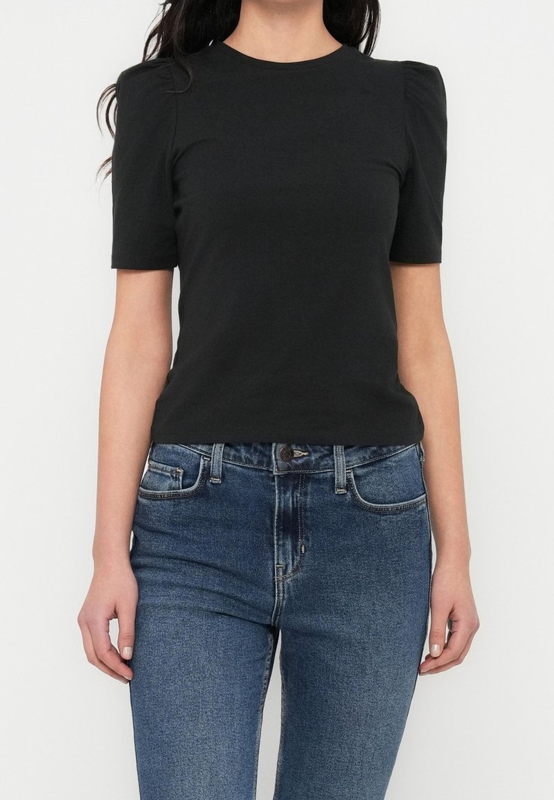 Femme vêtue d'un haut noir à manches courtes avec des épaules bouffantes et un jean bleu taille haute, sur fond blanc uni.