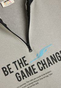 Sudadera gris con cremallera y cuello negro, que presenta un texto negro llamativo y un gráfico azul claro. Textura suave, diseñada para niños con un estilo deportivo.