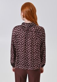 Blusa marrone a pois con collo arricciato e maniche lunghe. Il tessuto leggero presenta pois di colore crema di dimensioni alternate, grandi e piccoli.