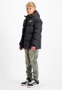 Zwarte pufferjack met een capuchon, voorzien van een logo op de borst en een ritsluiting. Gemaakt in combinatie met versleten lichtgroene jeans en zwarte sneakers.