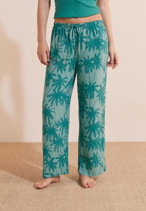 Personne portant un pantalon ample bleu sarcelle avec des motifs de palmiers bleu sarcelle foncé, debout pieds nus sur un sol texturé.