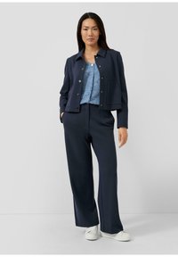 Marineblauwe cropped jas met zilveren knopen, gecombineerd met een lichtblauw textuurblouse en marineblauwe broek met wijde pijpen, afgewerkt met witte sneakers.