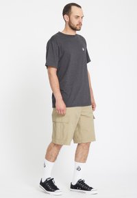 Camiseta de algodón gris, pantalones cortos de cargo beige con bolsillos laterales, zapatillas negras y calcetines blancos, de pie sobre un fondo blanco liso.
