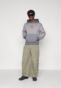 Sweatshirt cinza com gradiente e bolso frontal, calças cargo bege claro com zíperes e sapatos pretos. O modelo usa óculos de sol e está em uma pose relaxada.