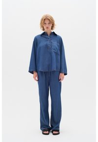 Denimový outfit skládající se z volné dlouhé košile s kapsou na hrudi a kalhot s volným střihem, obojí v středně modrém tónu.