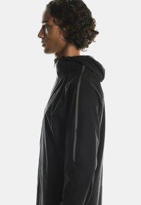 Giacca nera con cappuccio, caratterizzata da un design aerodinamico e strisce riflettenti lungo le spalle. Realizzata in un materiale leggero e texture.