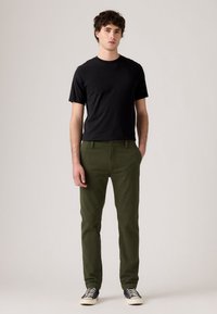 Man wearing black t-shirt, olive green pants, and black sneakers with white soles, standing with one hand in pocket against plain background.