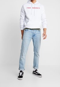 Vit hoodie med röd text "FAKE ROMANCE", ljusblå jeans och svarta Vans-sneakers. Hoodien har en känguruficka och dragsnoddar.