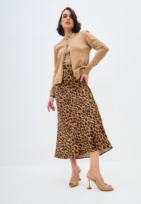 Vaaleanruskea neuletakki beige-paidan päällä, yhdistettynä leopardikuosisella midi-hameella ruskean ja kultaisen sävyissä. Beigen korkokengät täydentävät asua.
