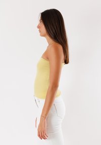 Top giallo senza spalline con una leggera lucentezza, abbinato a jeans bianchi aderenti. La modella ha i capelli lunghi e lisci ed è catturata di profilo.