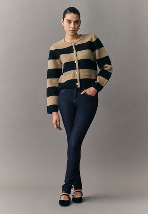 Žena oblečená v béžovom a čiernom pruhovanom kardigáne na gombíky, tmavé úzke džínsy, čierne baleríny, stojaca s jednou rukou vrecku pred jednoduchým pozadím.