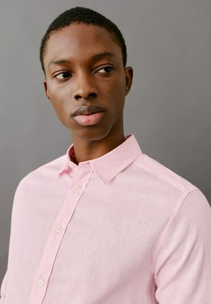 Giovane uomo con una camicia rosa chiaro con bottoni, che guarda leggermente di lato su uno sfondo grigio uniforme.