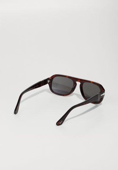 Óculos de sol em tartaruga castanha com armação quadrada, lentes escuras e acabamentos em metal nas dobradiças. Braços das laterais finos e curvos.
