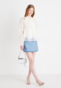 Gjennomsiktig hvit bluse med volanger og sløyfe, parret med en lys blå denimskjørt og sølvfarget veske; modellen har på seg sandalene med stropper.