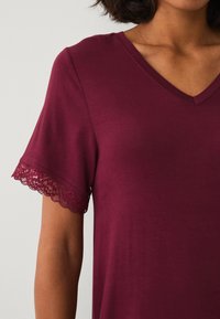 Maglietta bordeaux a scollo a V con maniche corte, caratterizzata da una delicata bordatura in pizzo sull'orlo delle maniche. Tessuto morbido con una texture liscia.