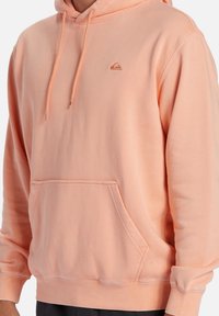 Sudadera color durazno claro con bolsillo frontal, cordones y un pequeño logo bordado en el pecho, llevada por una persona recortada a la altura de los hombros.