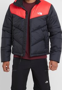 Giacca a vento nera con parte superiore rossa, cuciture orizzontali, colletto alto, chiusura con zip e tasche laterali. Logo del marchio sul petto.