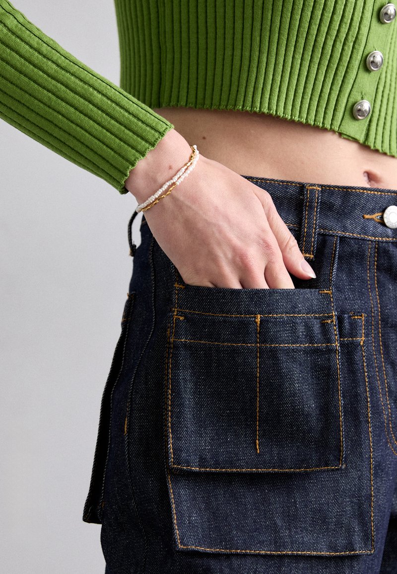 Crop top côtelé vert avec des boutons argentés, associé à un pantalon en denim noir taille haute avec des coutures orange et une poche arrière.