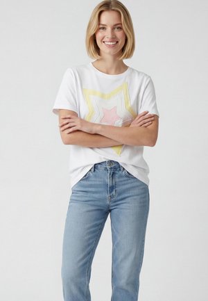 Donna sorridente con le braccia incrociate, indossa una maglietta bianca con una stella rosa e gialla e jeans azzurri su sfondo uniforme.