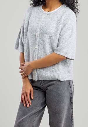 Vrouw die een lichtgrijs, kort mouwtjes, knoopjesvestje draagt en hoge taille, vervaagde zwarte jeans, staand met gekruiste handen.