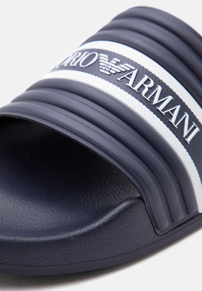 Κοντινό πλάνο ενός σαγιοναριού σε ναυτικό μπλε χρώμα με υφή στο πέλμα και λευκό-ναυτικό μπλε λουράκι που φέρει το λογότυπο Emporio Armani και το έμβλημα με τον αετό.