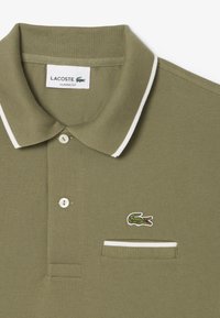 Polo verde oliva con colletto a coste, dettagli bianchi, due bottoni e un piccolo logo di coccodrillo ricamato sulla tasca del petto.