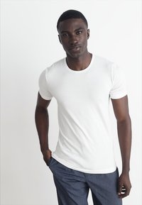 T-shirt blanc à manches courtes avec un col rond, en tissu lisse. Porté avec un pantalon bleu, au fit décontracté.