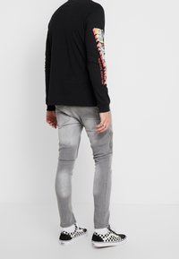 Camisa negra de manga larga con un gráfico colorido en el brazo izquierdo, combinada con pantalones gris claro y zapatillas de lona a rayas en blanco y negro.